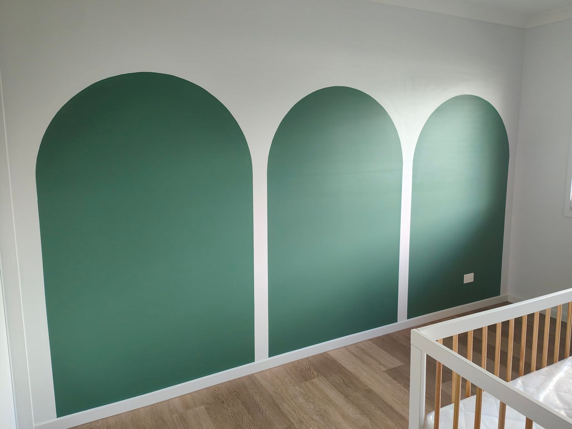 Three green arched wall panels painted on a white nursery wall above wood flooring