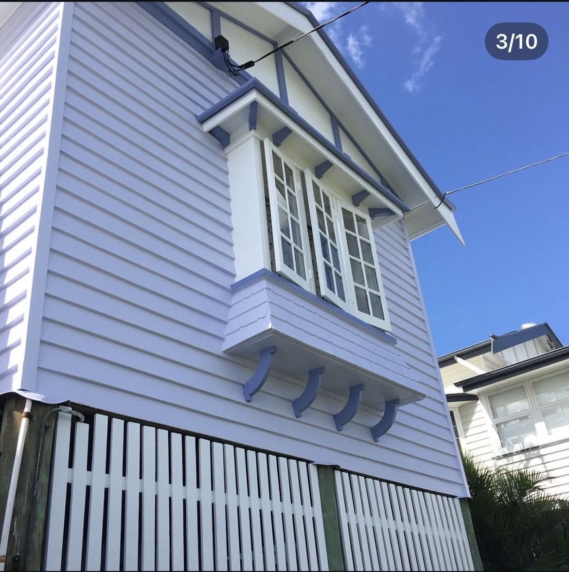 Light purple house exterior with white-trimmed windows, decorative brackets, and a slatted fence under a blue sky