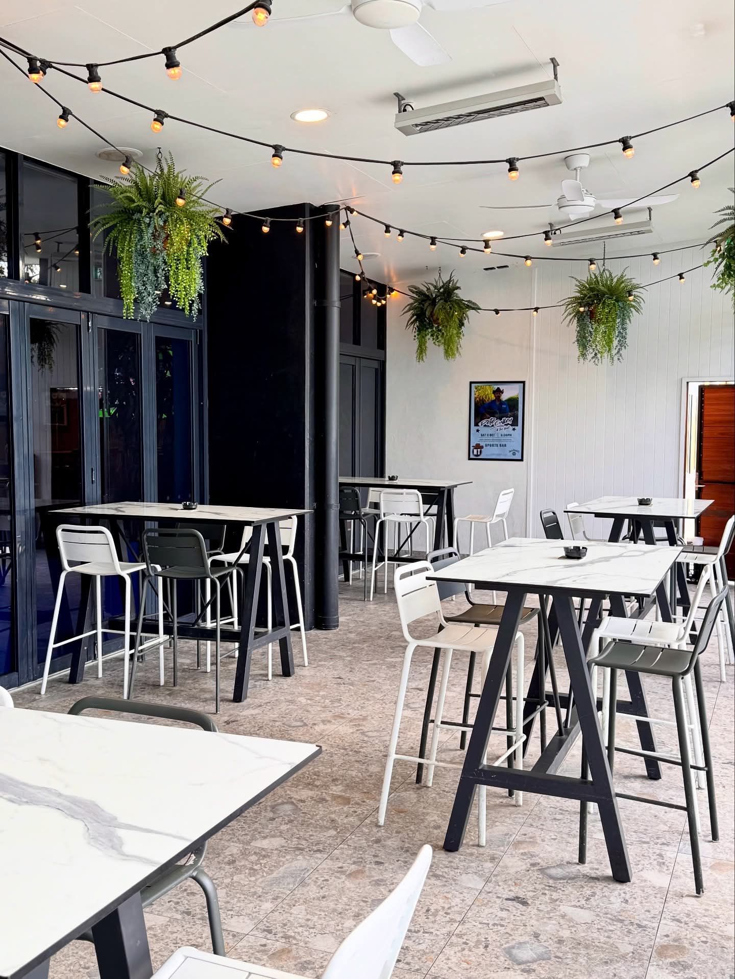 Bright café patio with white tables, black stools, hanging string lights, and potted plants
