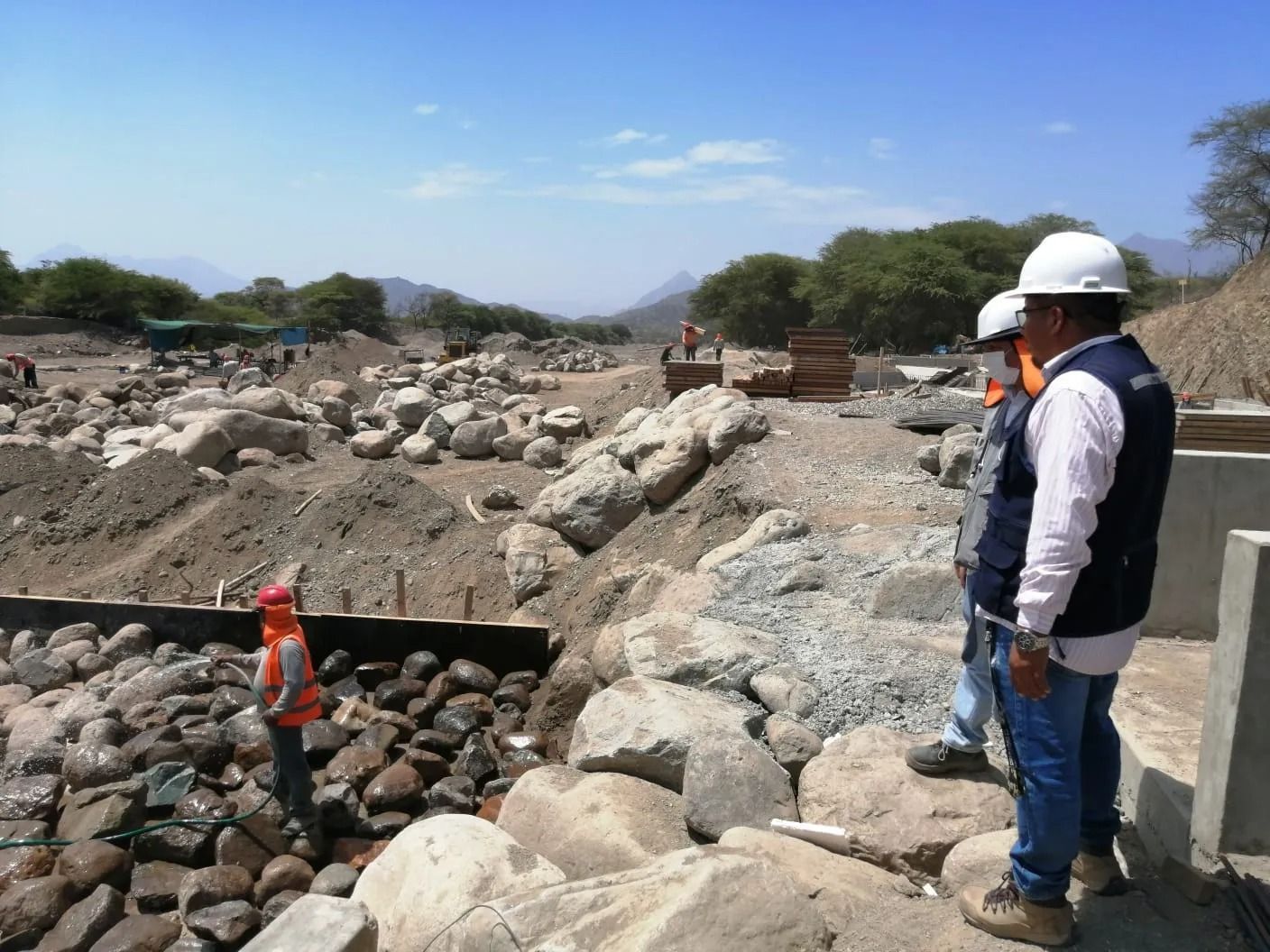 Dos obreros con cascos inspeccionan una obra rocosa junto a un estrecho canal.