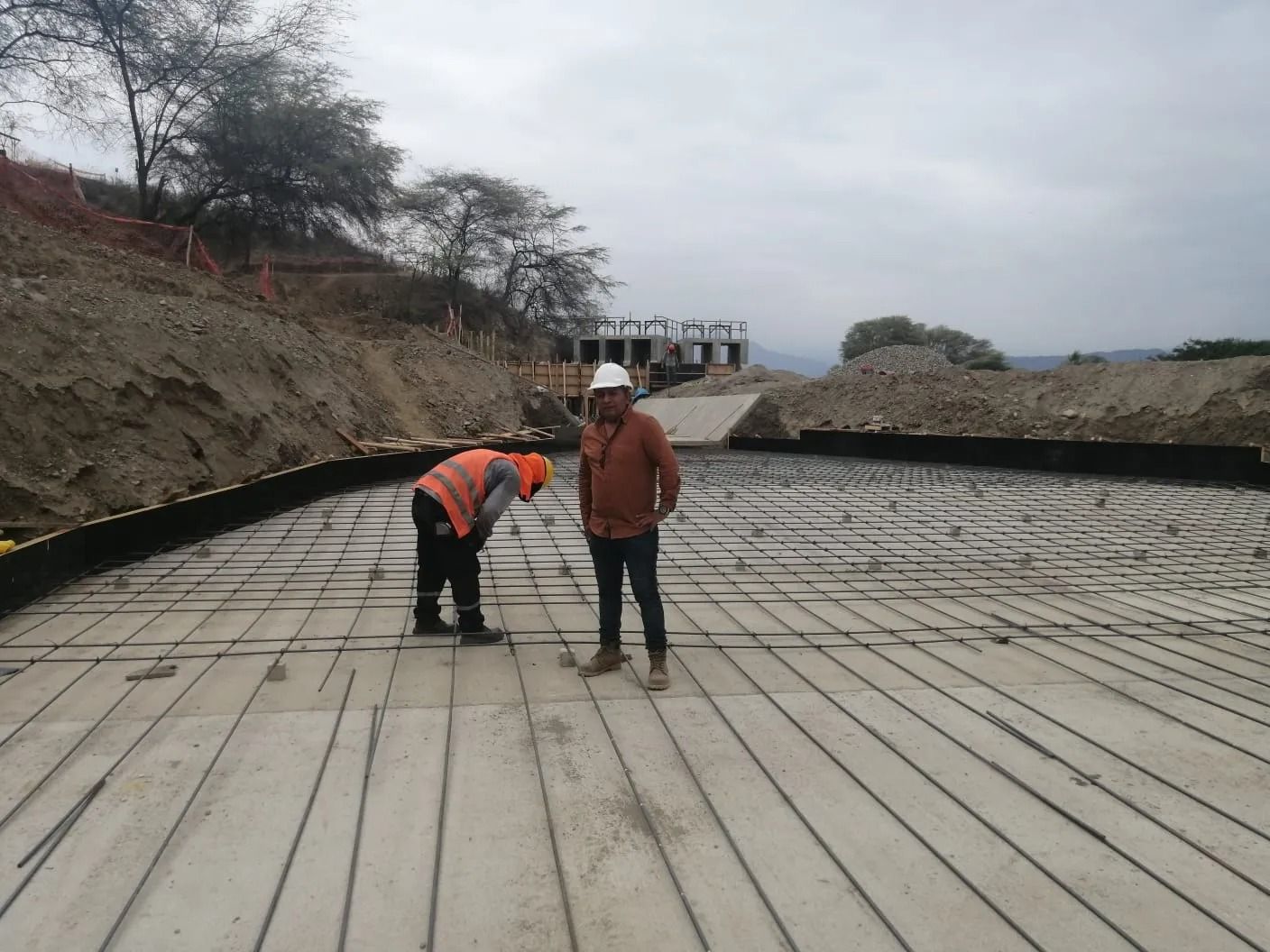Dos trabajadores sobre una losa de hormigón en una obra en construcción; uno está agachado y el otro permanece de pie cerca.