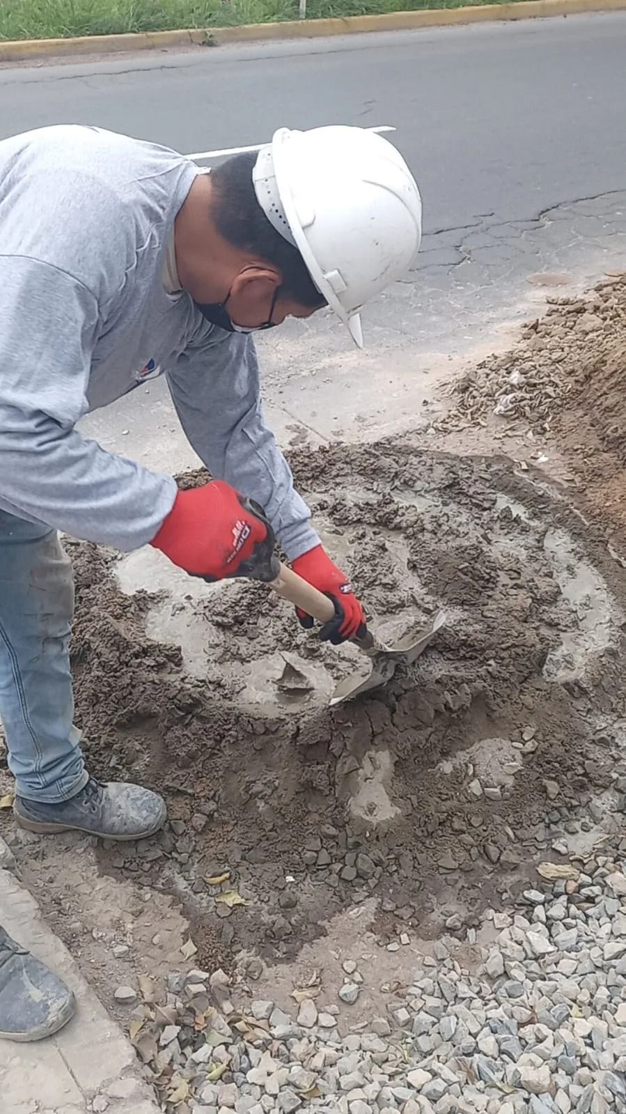 Un trabajador con casco y guantes rojos mezclando hormigón fresco junto a una carretera.