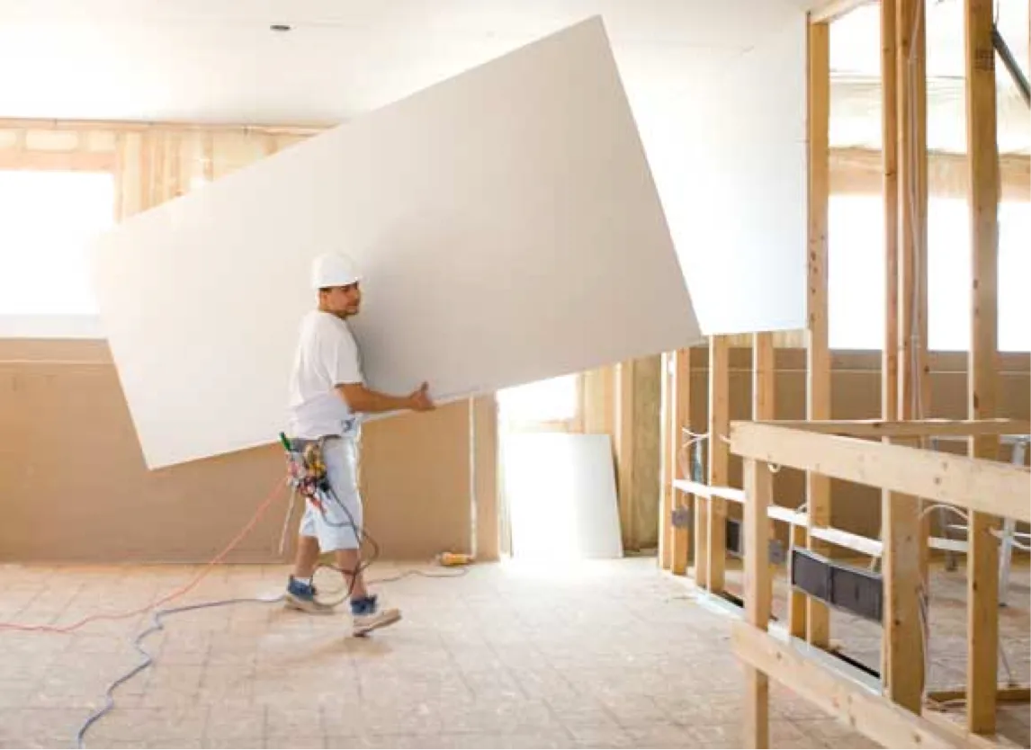 Un trabajador transporta una gran lámina de paneles de yeso dentro de una habitación en construcción.