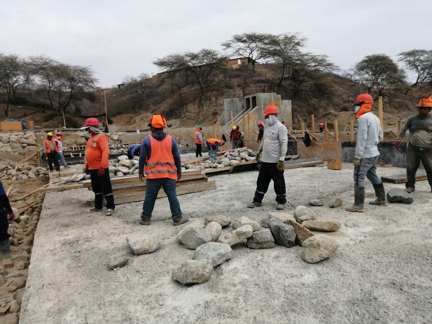 Obreros de la construcción con chalecos naranjas y cascos en un sitio de construcción rocoso con materiales dispersos.