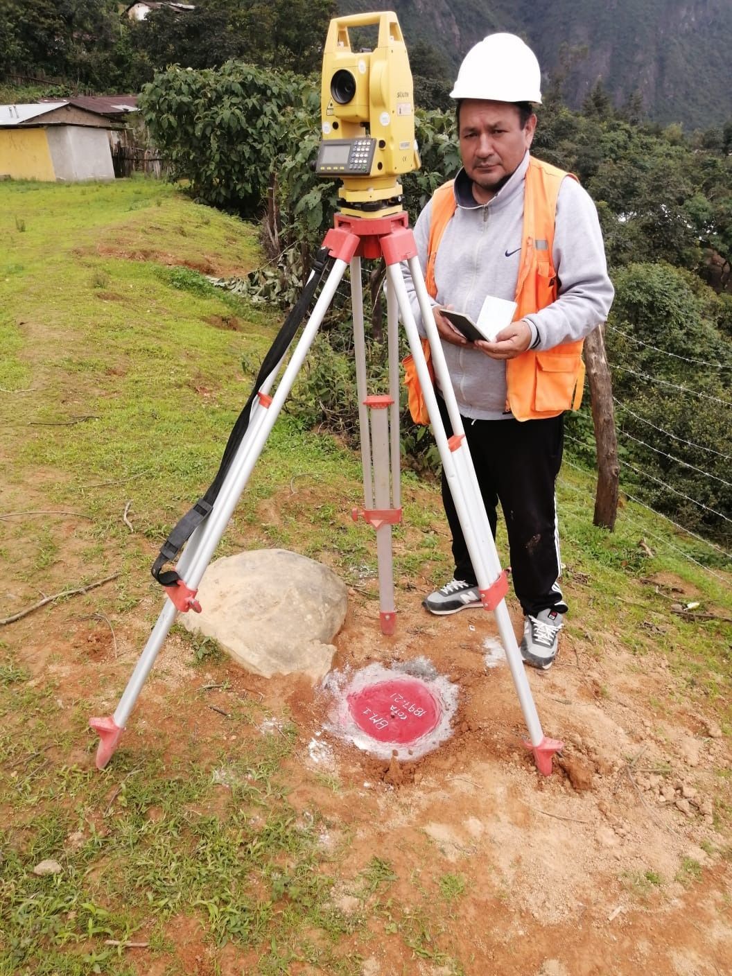 Topógrafo con casco utilizando un teodolito montado sobre trípode en un terreno cubierto de hierba.