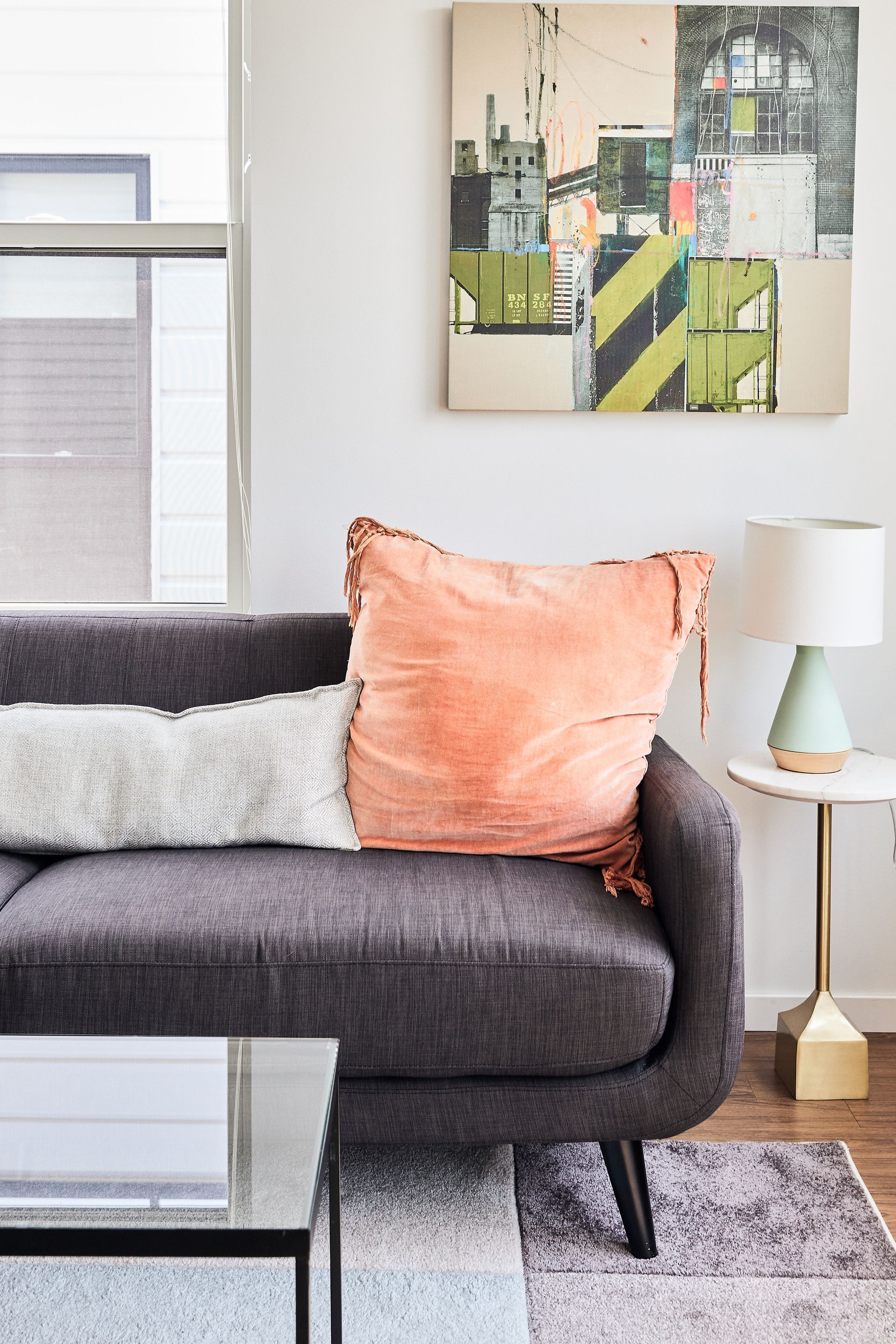 Gray couch with coral pillow, white pillow, painting, side table, and lamp at Southsider Apartments in Minneapolis, MN.