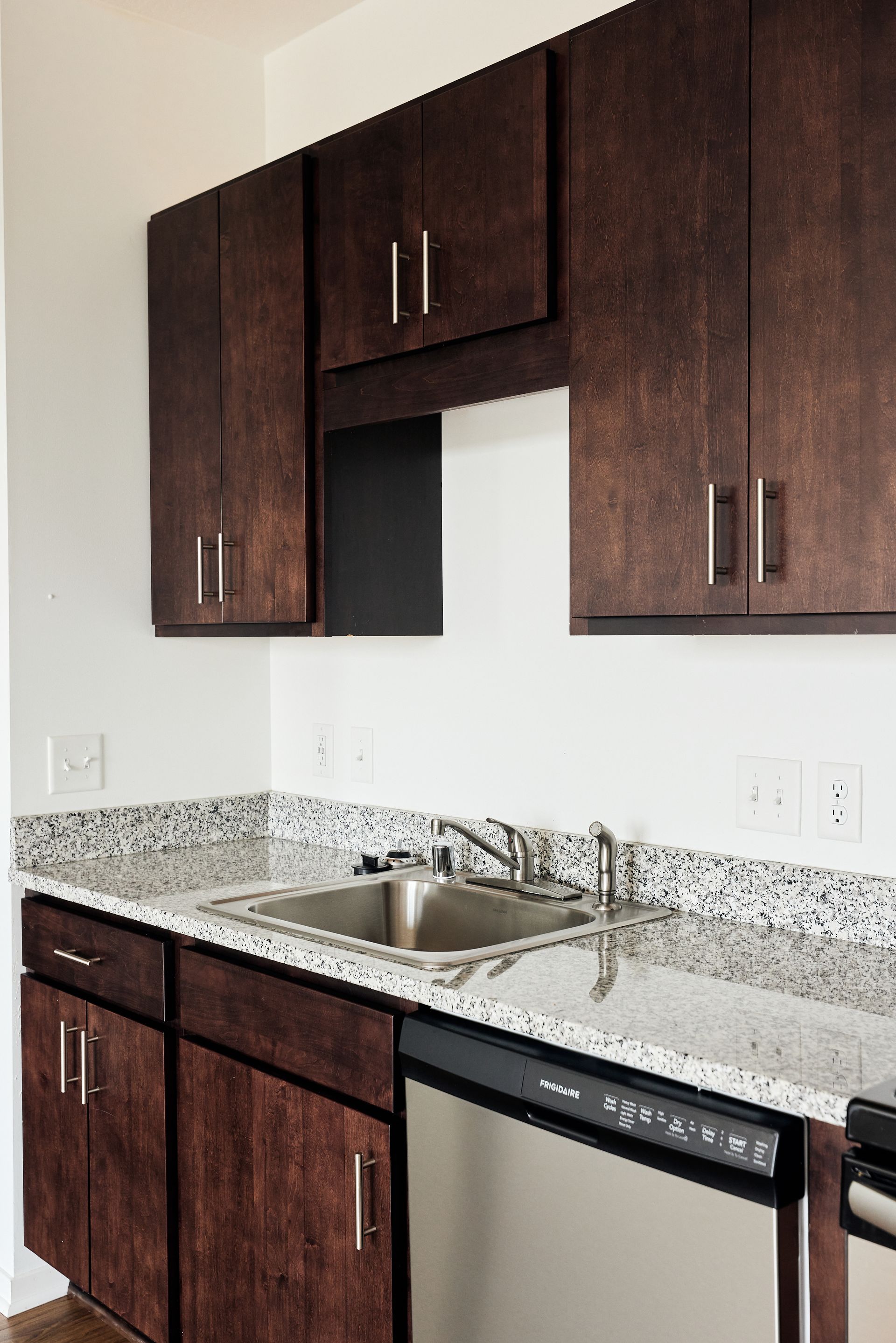Dark brown kitchen cabinets above a stainless steel sink and dishwasher; granite countertop at Southsider Apartments in Minneapolis, MN.