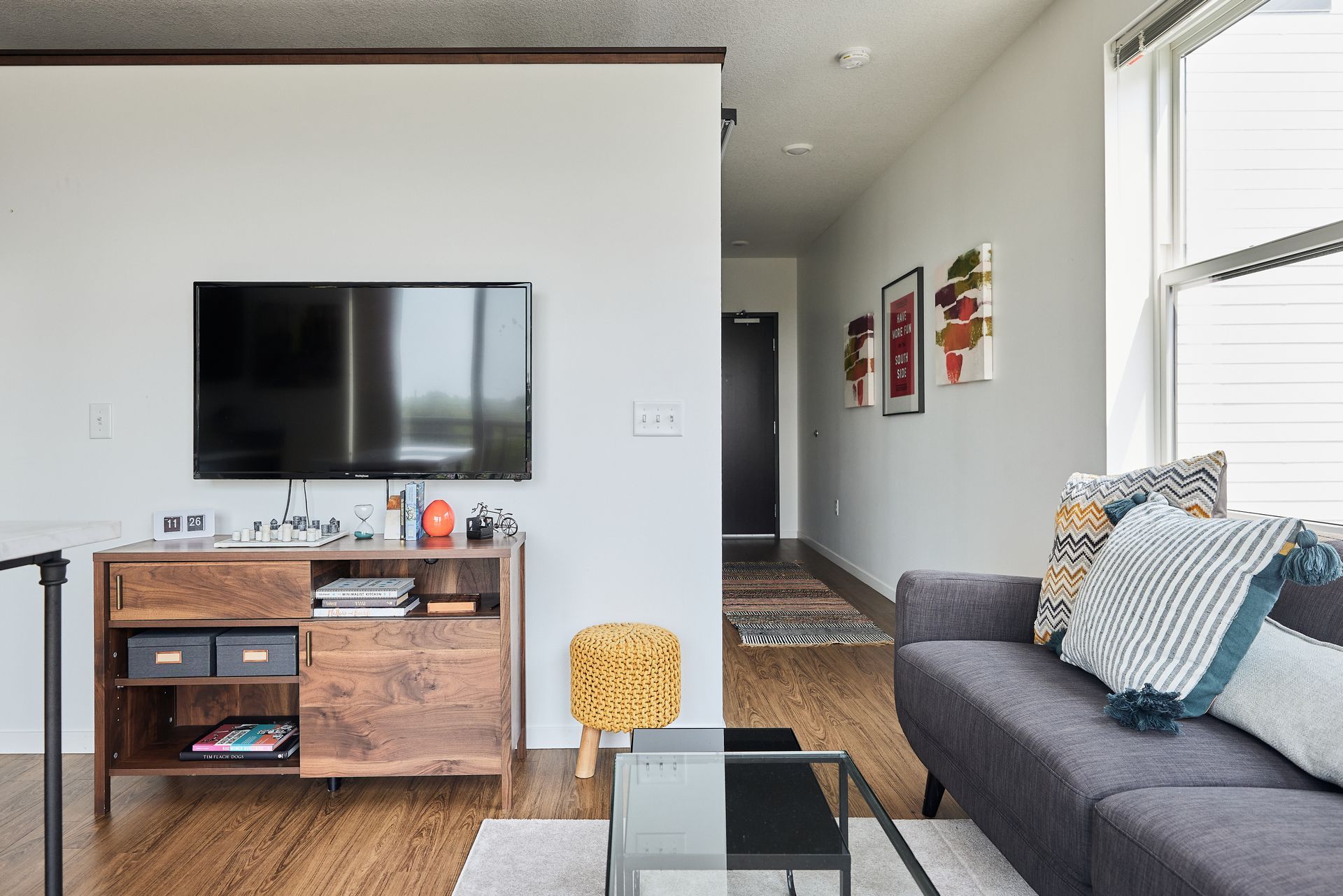 Living room with TV, wooden cabinet, gray couch, and hallway with artwork at Southsider Apartments in Minneapolis, MN.