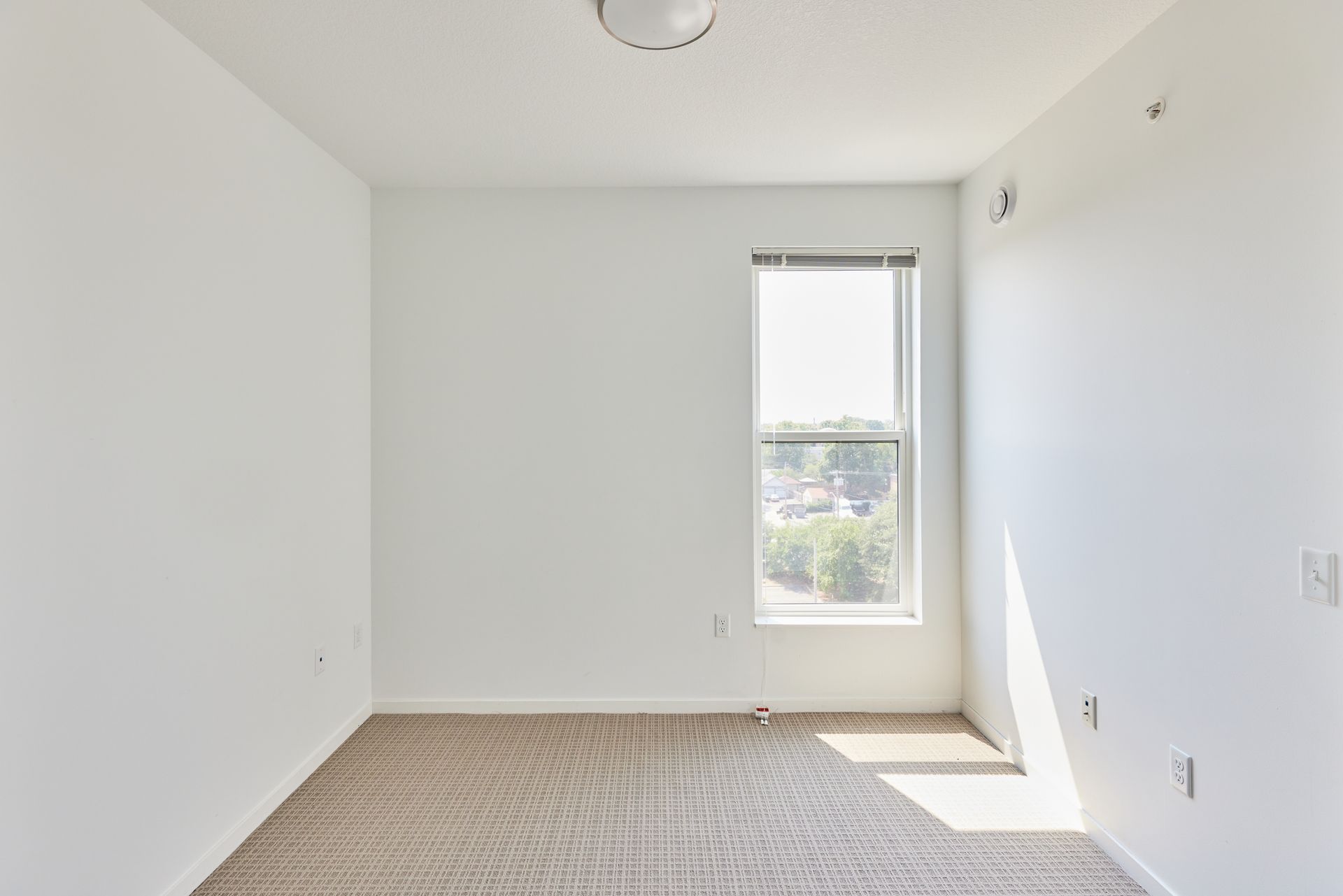 Empty room with beige carpet, a small window, and white walls at Southsider Apartments in Minneapolis, MN.