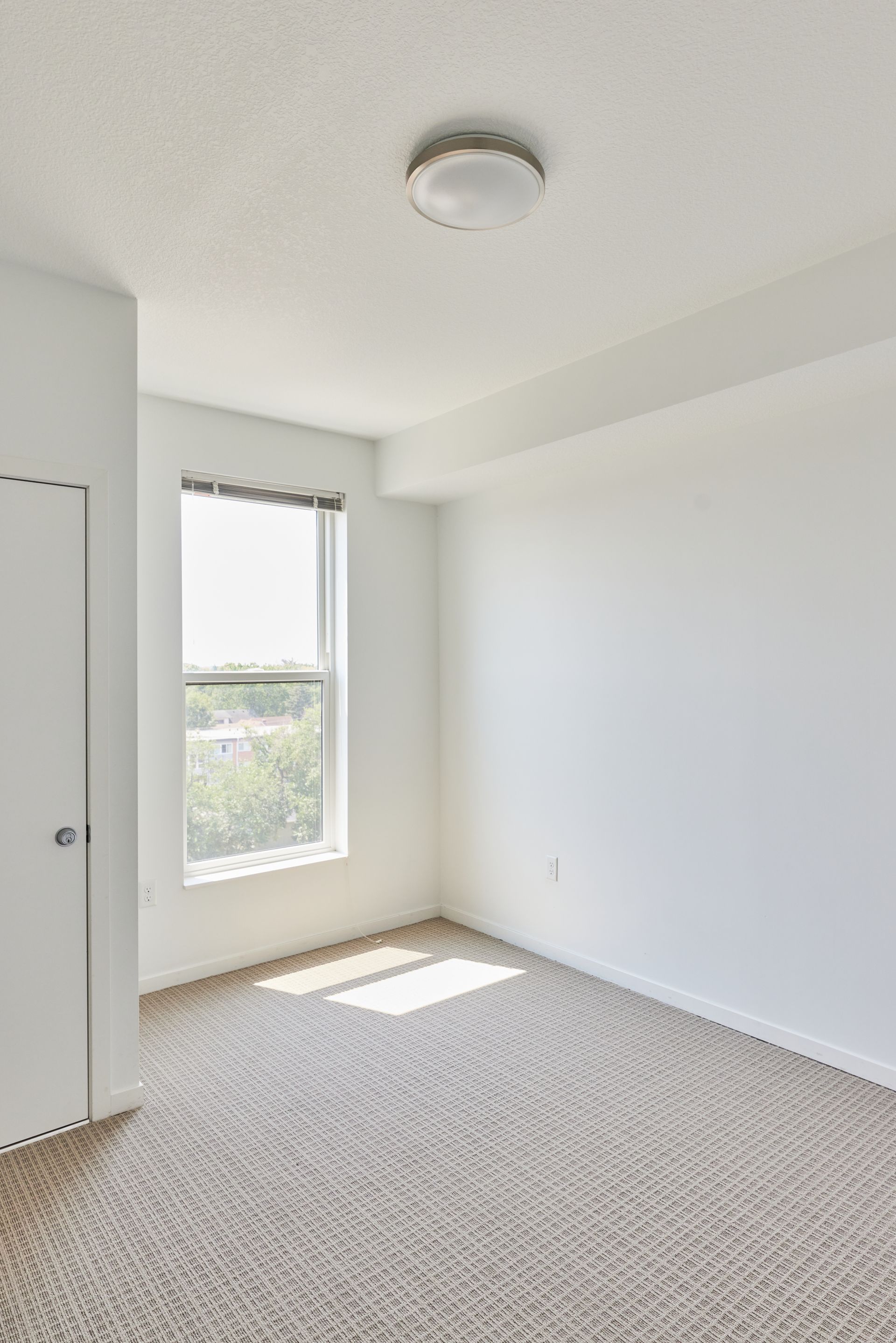 Empty room with beige carpet, a window, and a closed white door at Southsider Apartments in Minneapolis, MN.