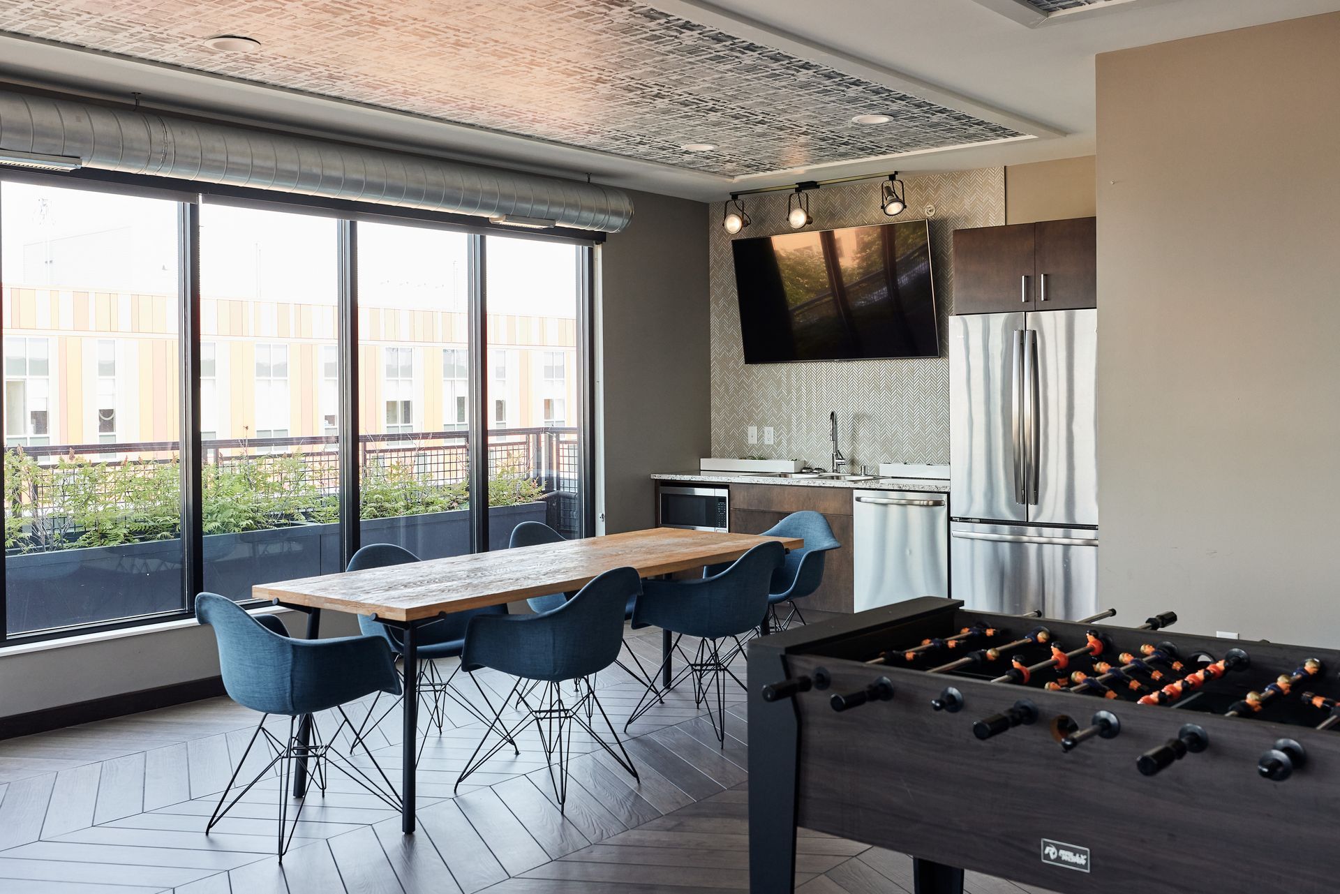 A modern lounge with a foosball table, dining table, kitchen, and a large window with a cityscape view at Southsider Apartments in Minneapolis, MN.