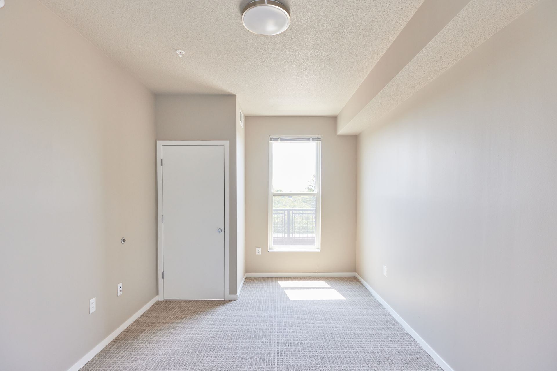 Empty beige-walled room with a window, white door, and light carpet at Southsider Apartments in Minneapolis, MN.