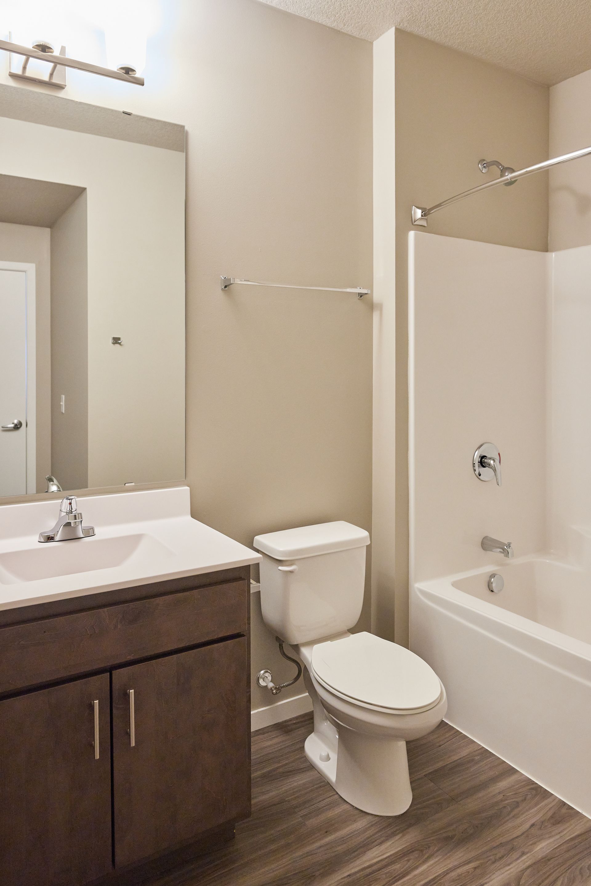 Bathroom with white tub, toilet, and sink. Brown vanity and light gray walls, with wooden floor at Southsider Apartments in Minneapolis, MN.