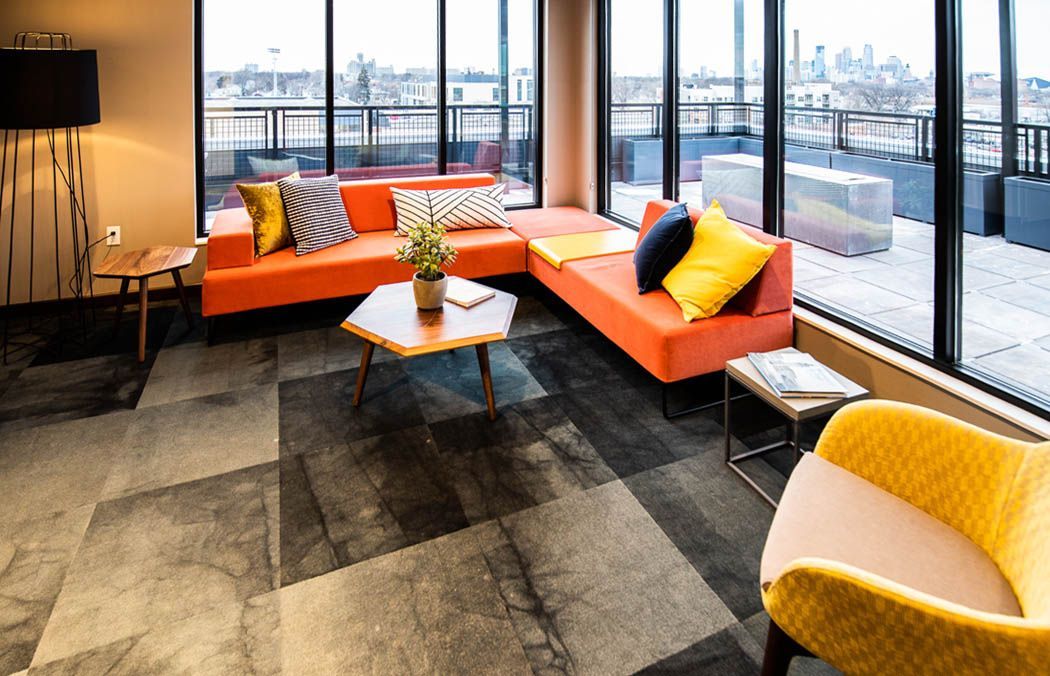 Modern lounge with orange sectional, yellow chair, patterned gray floors, large windows, and city view at Southsider Apartments in Minneapolis, MN.