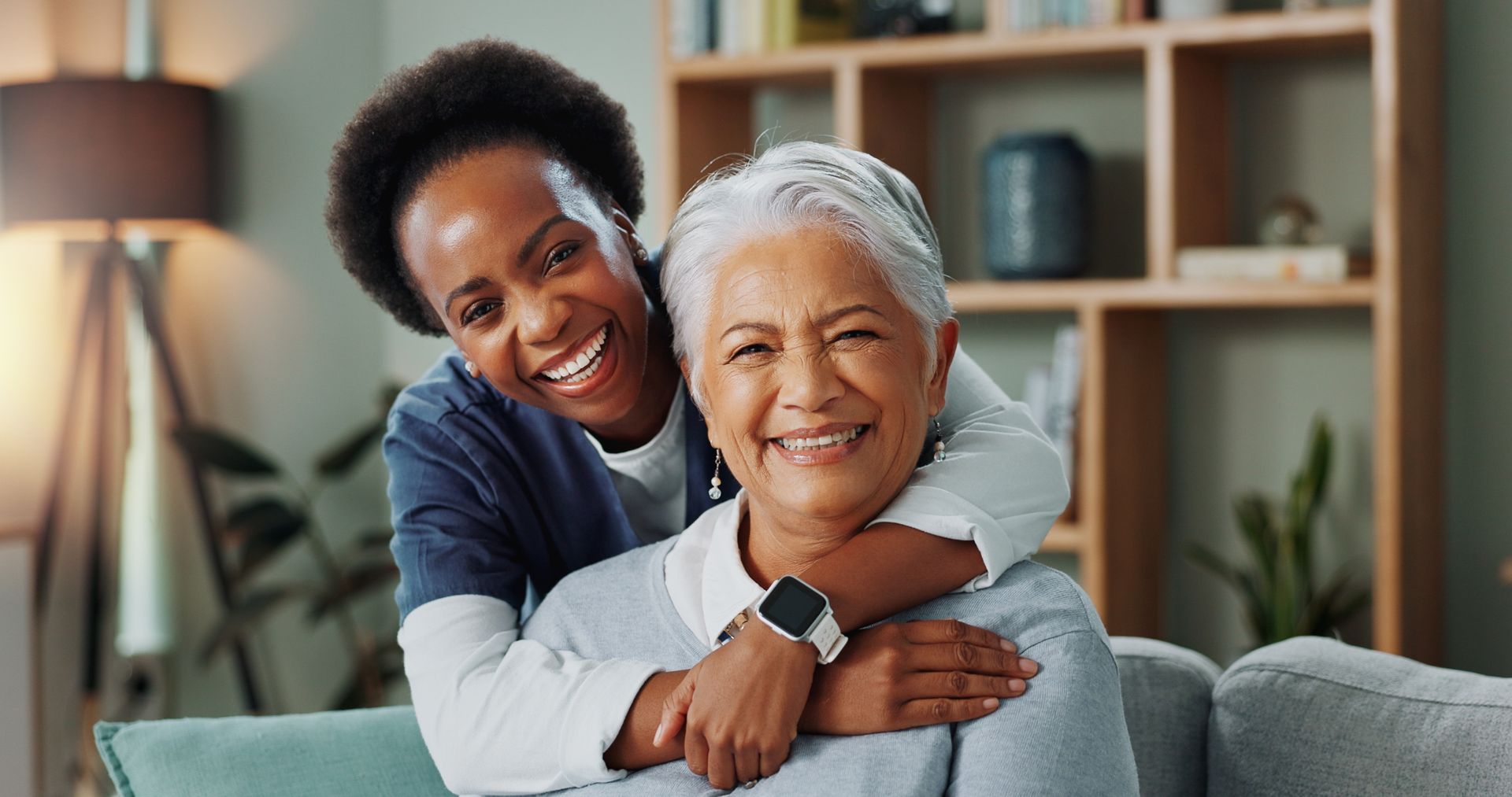 Two people smiling on a couch, with one hugging the other in a cozy living room.