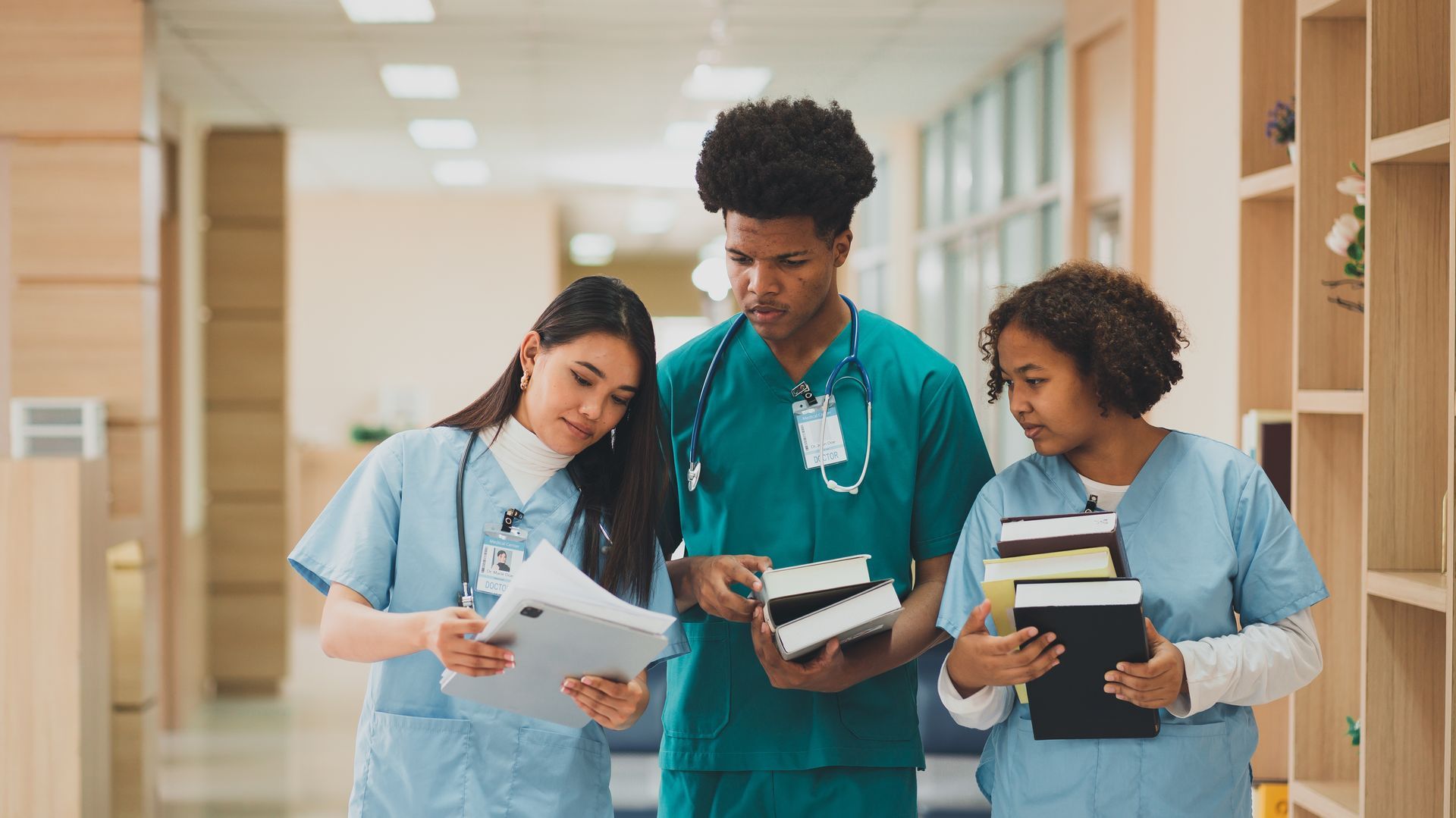 Three students in scrubs and coats discussing notebooks in a bright school hallway