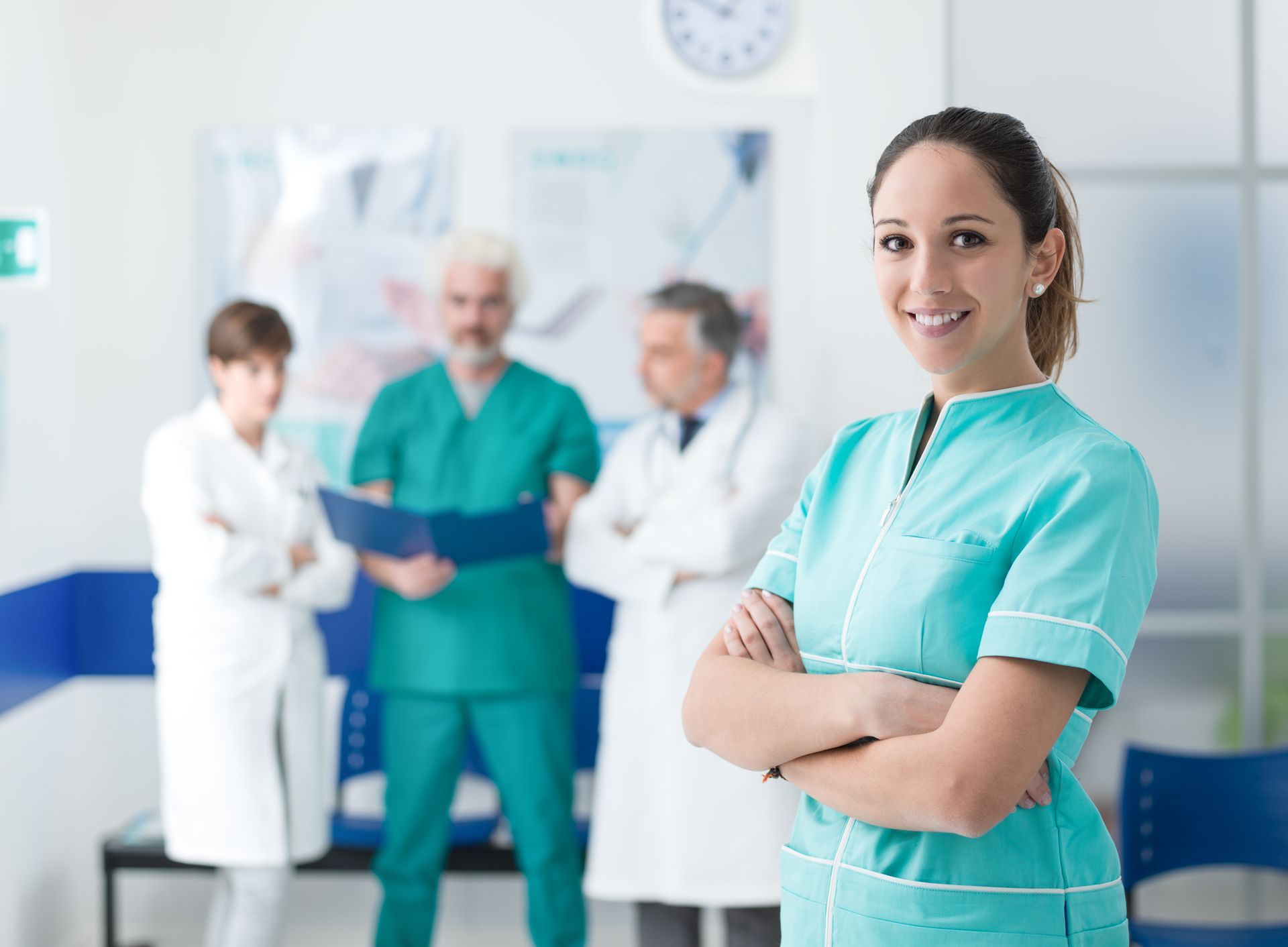 Nurse in teal scrubs smiling in a hospital hallway with doctors talking in the background