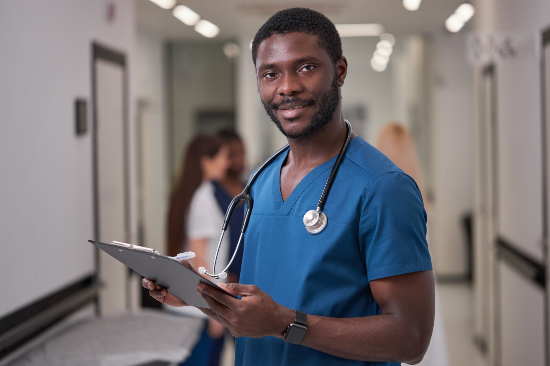 Nurse in blue scrubs holding a clipboard in a hospital hallway, smiling at the camera