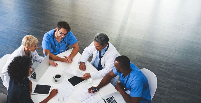 Four coworkers in blue and white shirts collaborate around a table with a tablet and coffee cups.