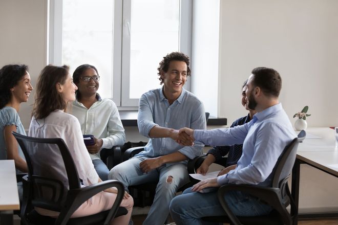 People seated in a meeting room, talking and smiling around a table near a bright window.
