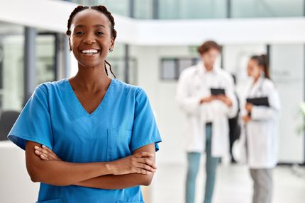 Smiling healthcare worker in blue scrubs with arms crossed, standing in a hospital hallway with doctors in the background