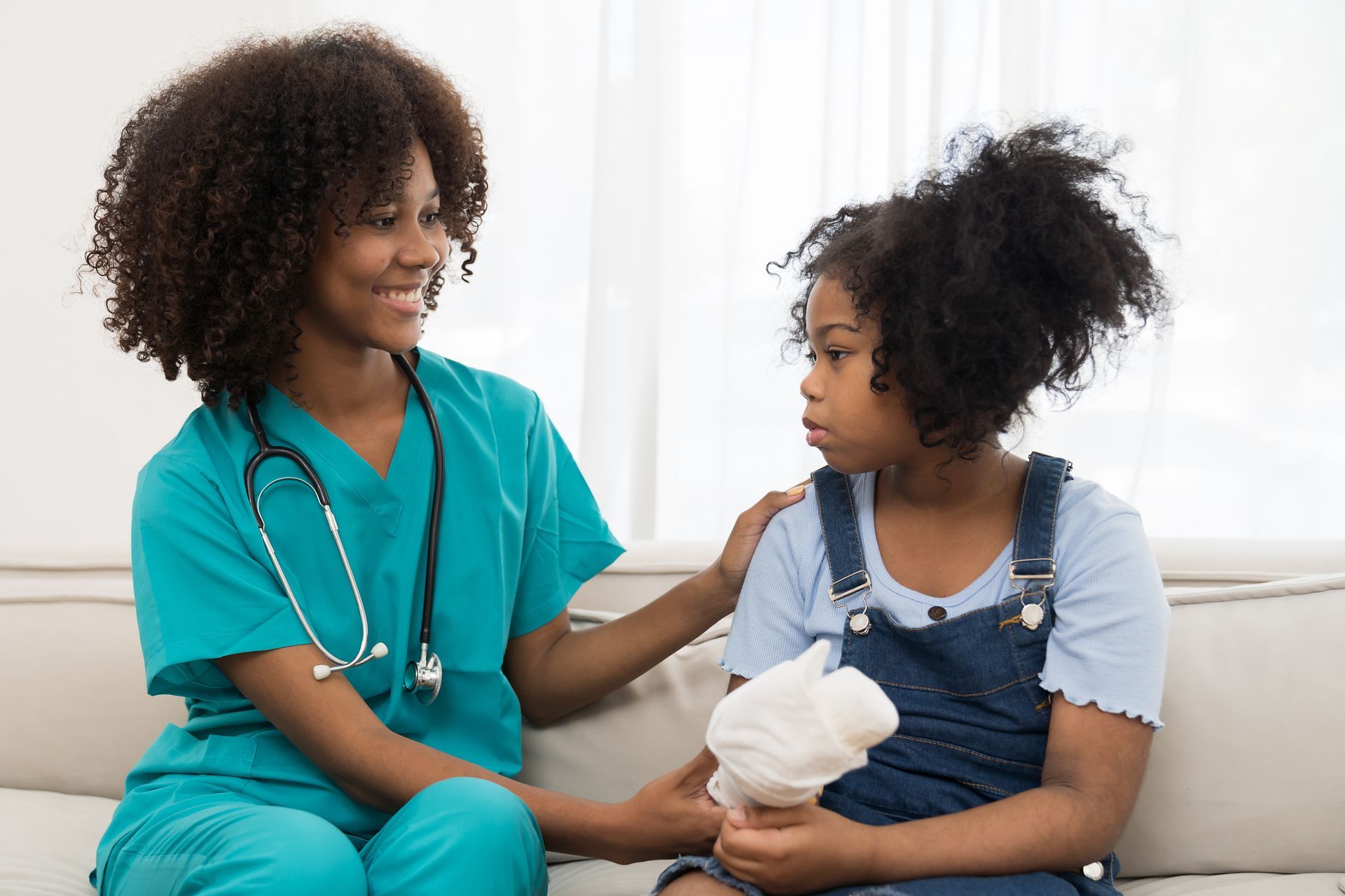 Nurse in teal scrubs talking with a patient on a couch, holding a tissue in a bright clinic setting