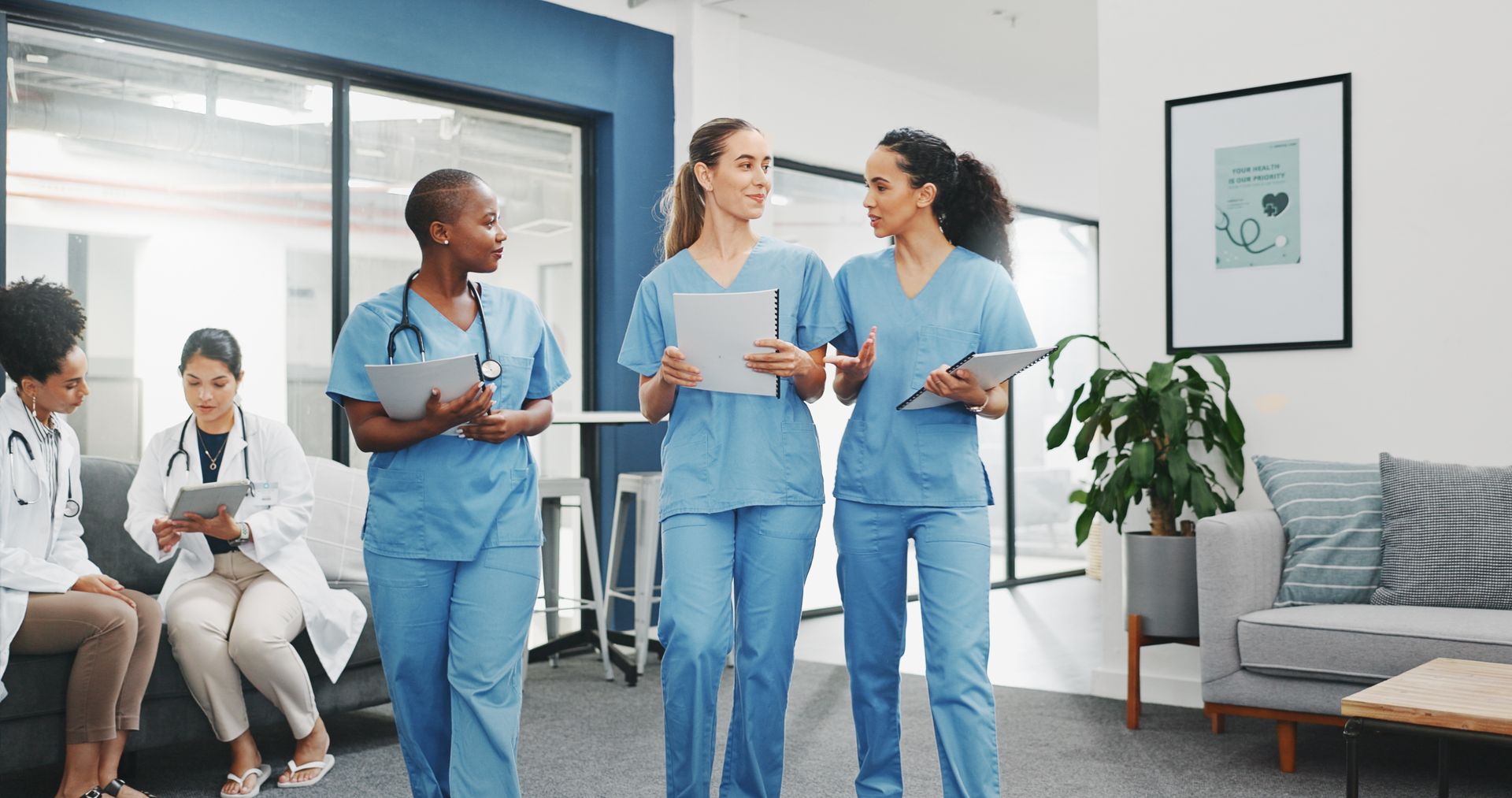 Hospital staff in blue scrubs talking in a bright waiting area beside seated patients