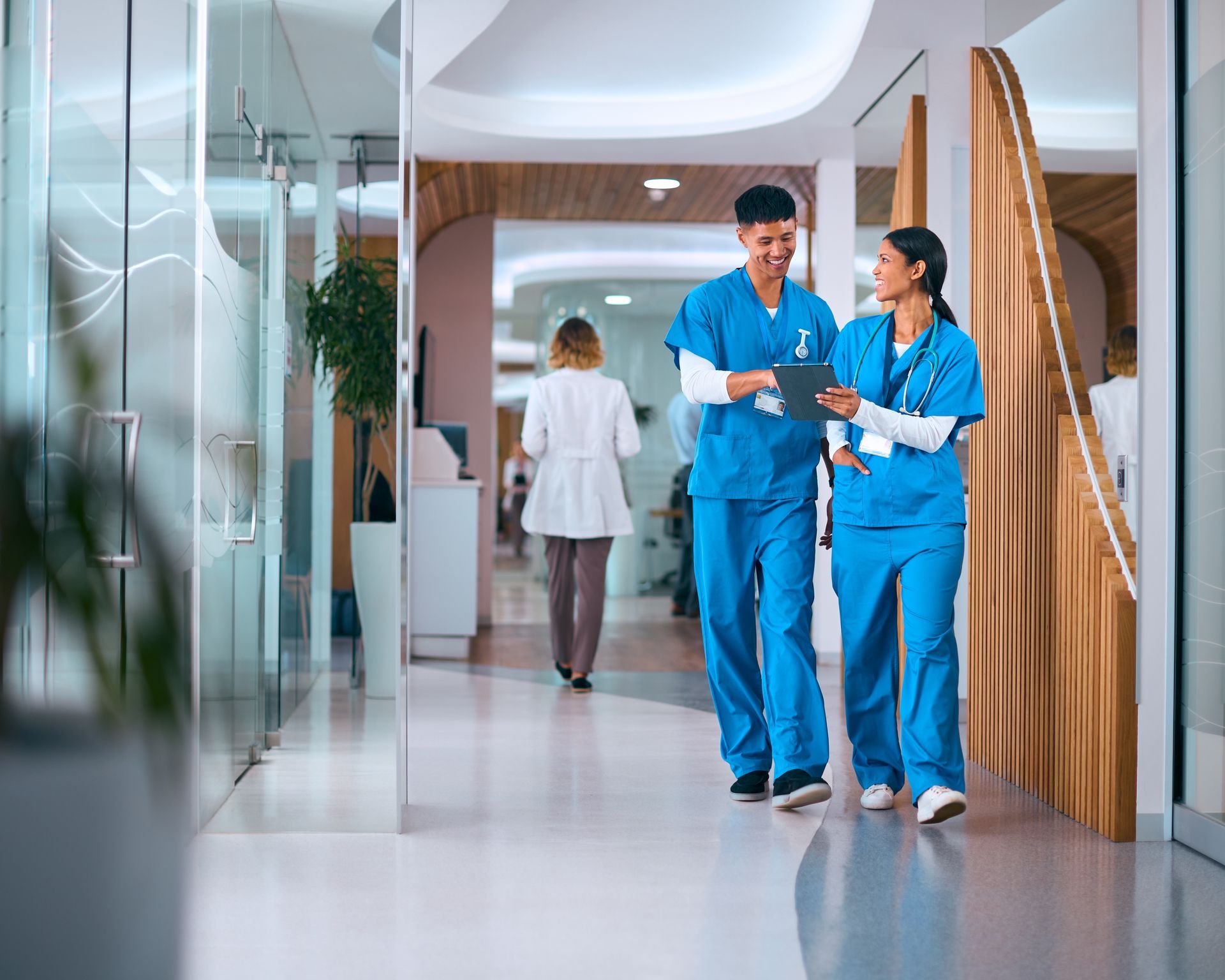 Two healthcare workers in blue scrubs walking and talking in a bright hospital corridor