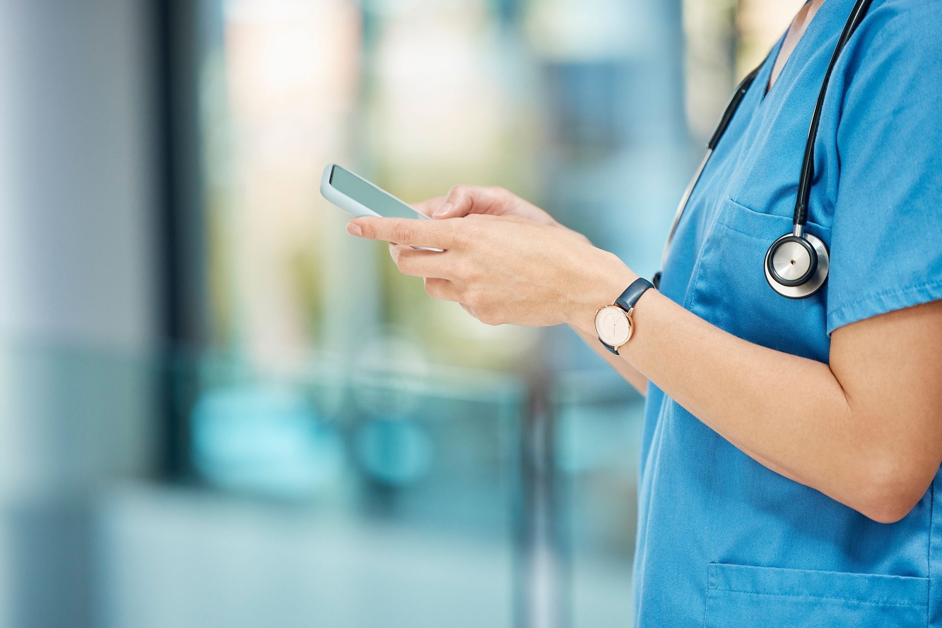 Healthcare worker in blue scrubs using a smartphone with a stethoscope around their neck