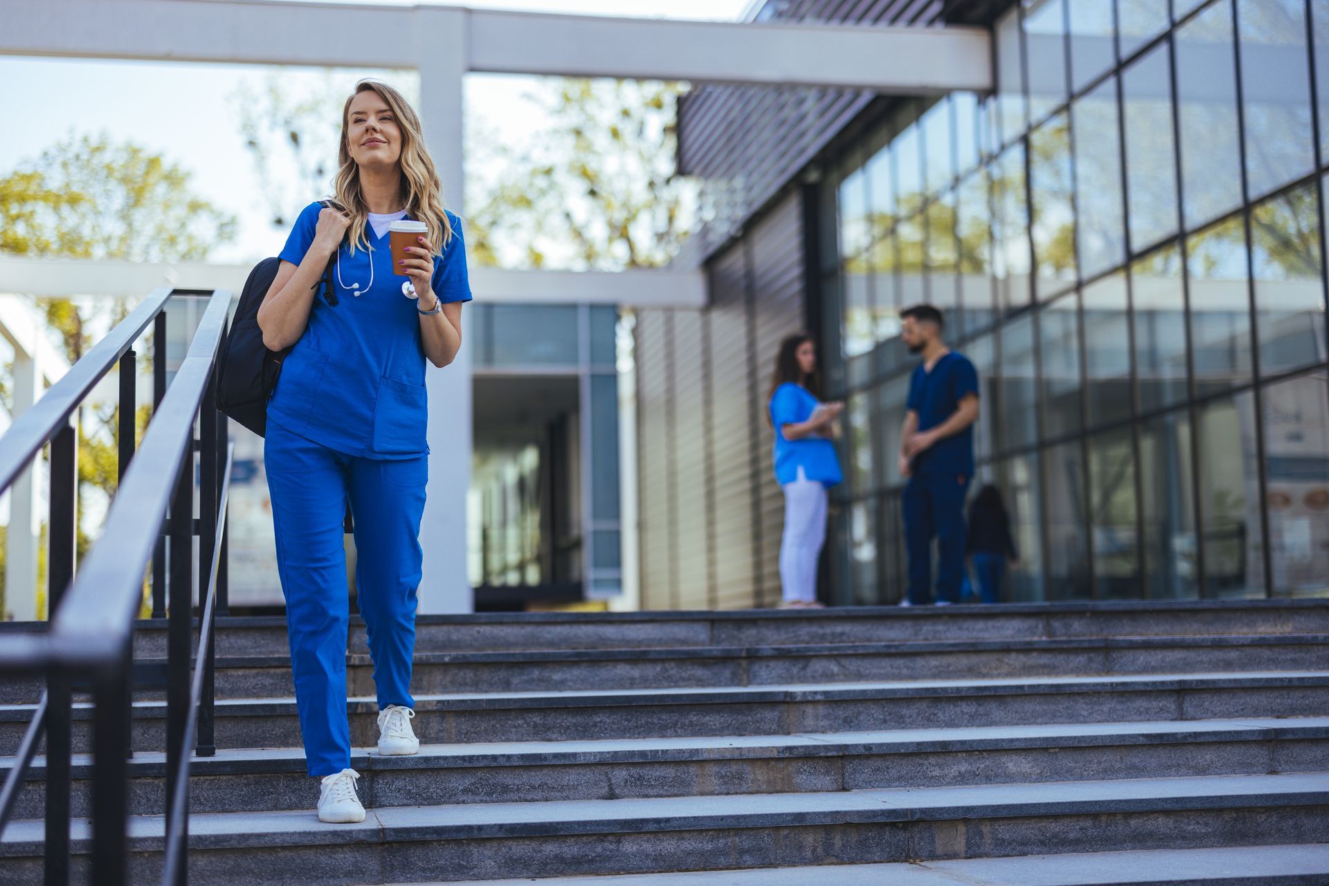 Woman in blue scrubs walking down outdoor steps with a coffee, beside a modern glass building
