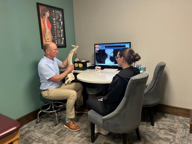 A professional in a blue shirt holds a spinal model while discussing a cervical X-ray with a patient at a desk.