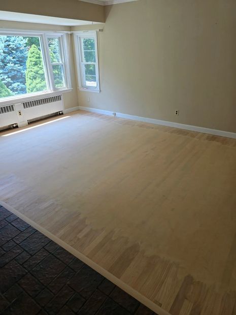 Empty beige living room with large windows and light wood floors, partially covered by dark carpet at left