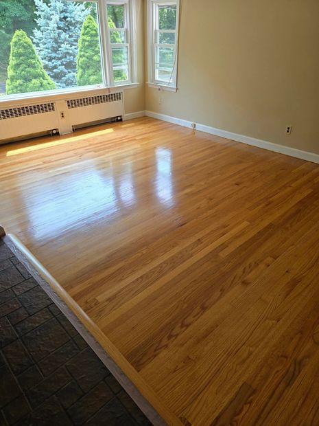 Empty sunlit room with polished hardwood floors, beige walls, and large windows overlooking trees