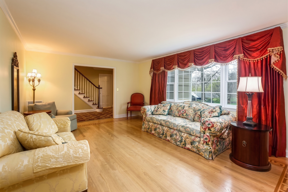 Bright living room with floral sofas, wood floors, red curtains, and a bay window