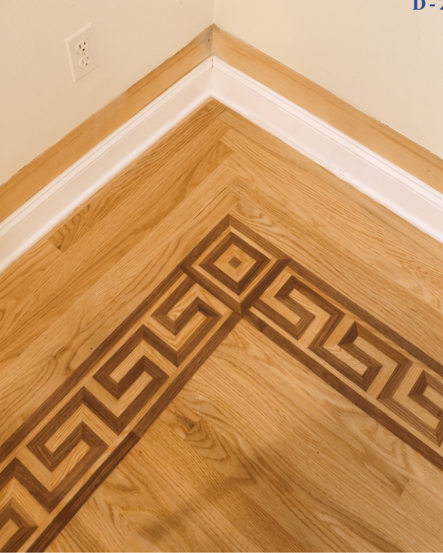 Corner of a wood floor with a dark geometric border pattern and white baseboard trim