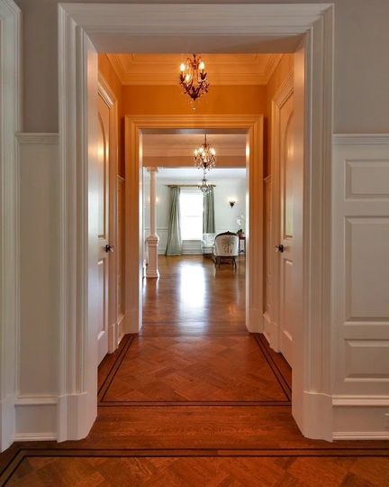 Elegant hallway with wooden floors, white trim, and chandeliers leading into a bright room
