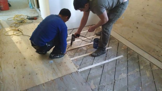 Two workers installing floor boards in a room, one kneeling and one bending over a cutout opening.