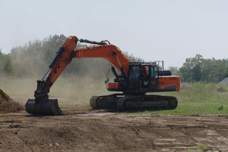 Orange excavator digging dirt on a construction site, with dust and trees in the background.