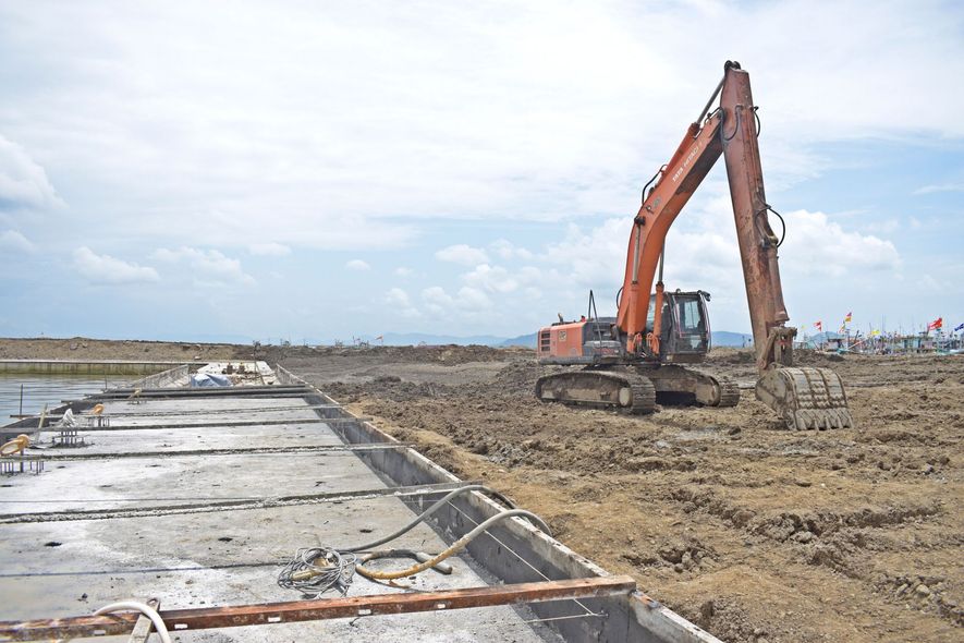 Orange excavator on a muddy construction site beside concrete slabs under a cloudy sky.
