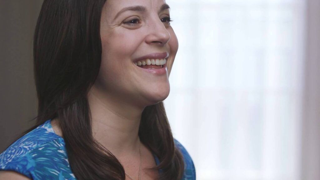 A woman in a blue dress is smiling in front of a window.