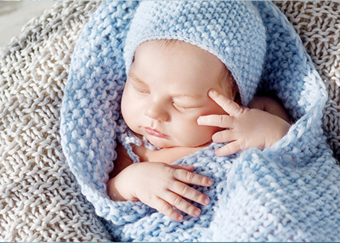 A newborn baby wrapped in a blue blanket and hat is sleeping in a basket