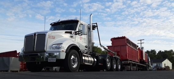 A white semi-truck with a roll-off trailer loaded with red shipping containers parked against a blue, cloudy sky.