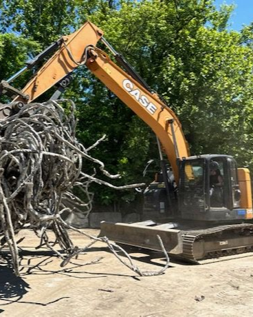 A yellow Case excavator uses a grapple attachment to lift a large, tangled mass of scrap metal outdoors.