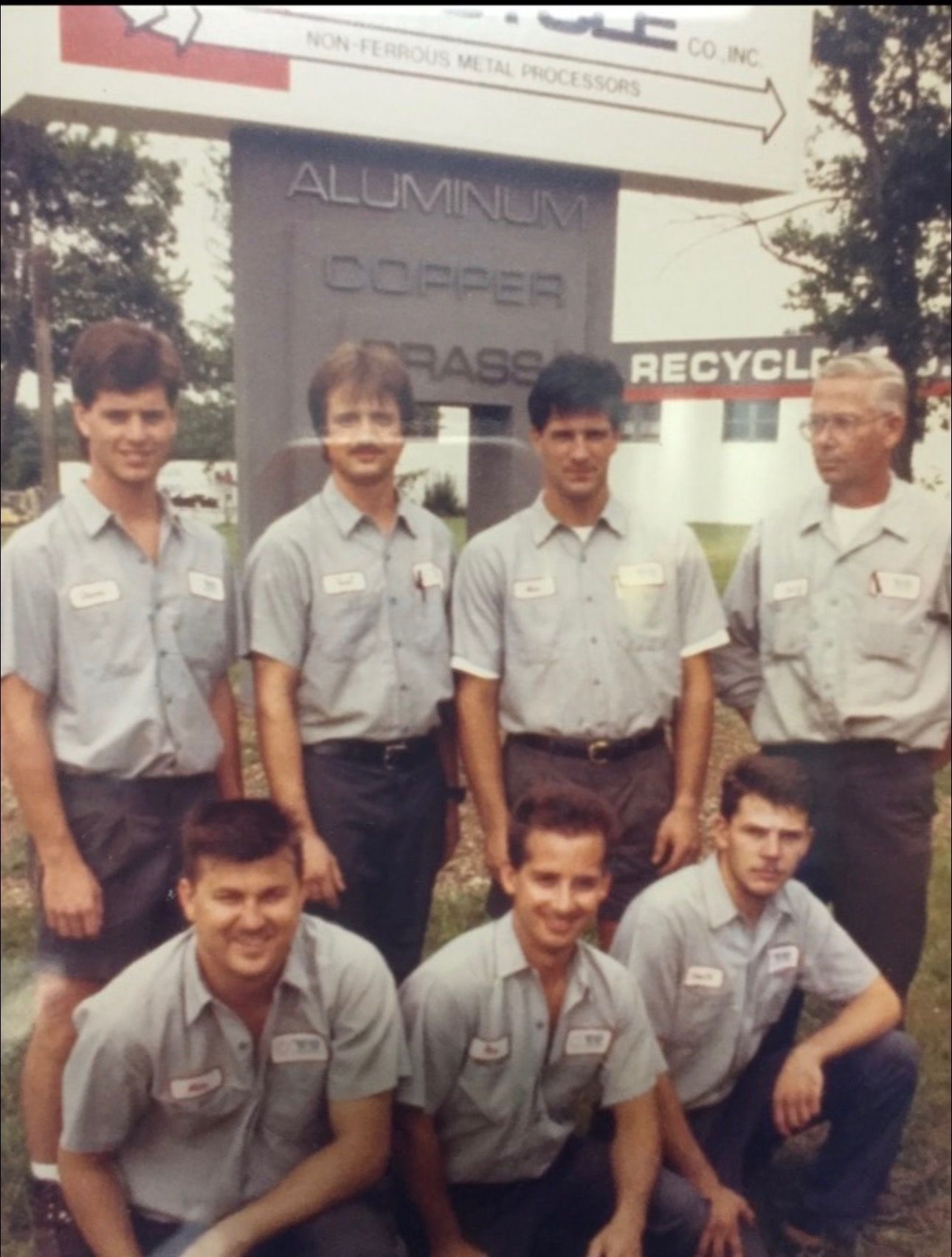 Seven staff in gray work uniforms pose outside a metal recycling facility with a sign reading Aluminum, Copper, Brass.