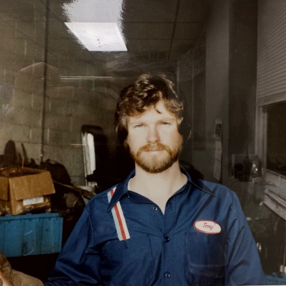 A person with brown, wavy hair and a beard wearing a blue work shirt with a name tag, standing inside a workshop.