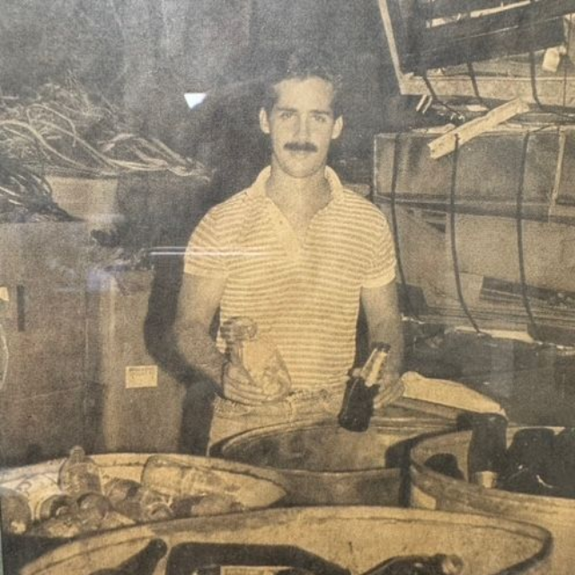 A man with a mustache in a striped polo shirt stands in a warehouse surrounded by bins of bottles.
