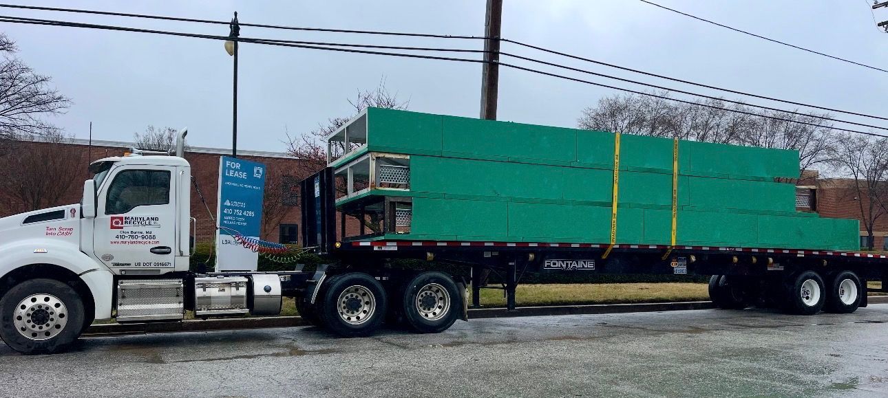 A white semi-truck hauls a flatbed trailer loaded with large, green-wrapped bundles of construction materials.