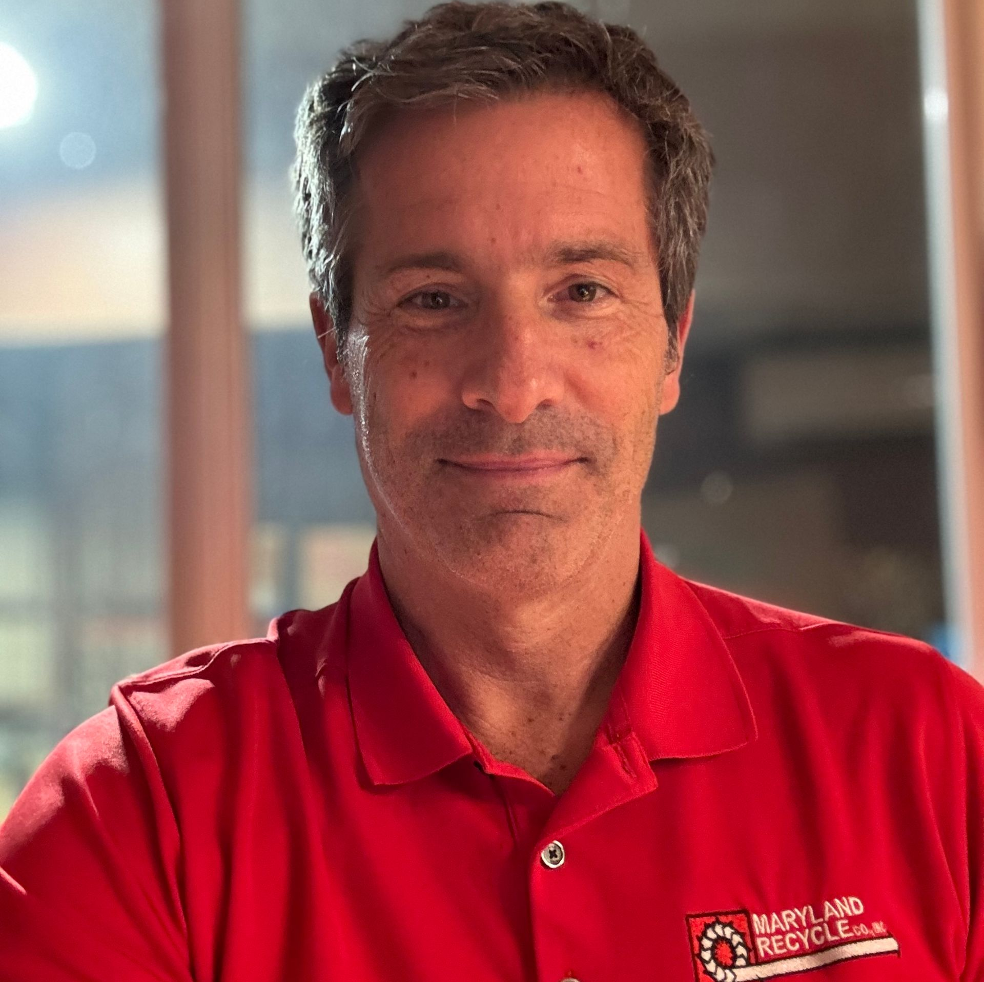 A smiling person in a red polo shirt with a Maryland Recycle logo, indoors against a blurred background.