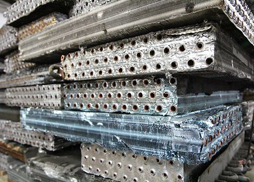 Stacked metal scaffolding planks with perforated surfaces, wrapped in plastic and piled in a storage area