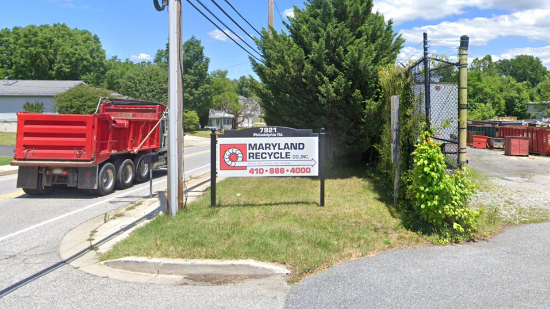 A red dump truck drives past a white sign for Maryland Paving on a grassy corner on a sunny day.